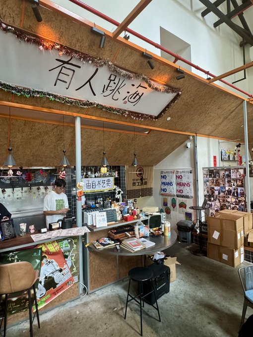 An interior view of a cafe or bar with Chinese signage reading '有人跳沙' hanging from the ceiling, featuring a counter, stools, a display of polaroid photos, and a staff member working behind the bar.