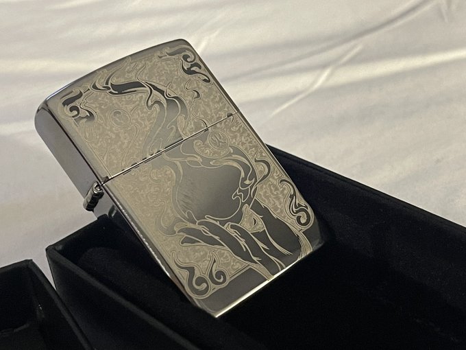 A polished silver Zippo-style lighter with ornate engraved tribal/floral designs sits in a black presentation box.