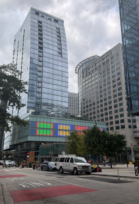 A modern glass high-rise with colorful panels on its lower floors towers over a city intersection, where a white van and silver car wait at a crosswalk marked "ONLY."

The image's urban rescue theme fits the post's playful "should I rescue @degentalks" caption, with the towering building serving as a dramatic "rescue" backdrop.