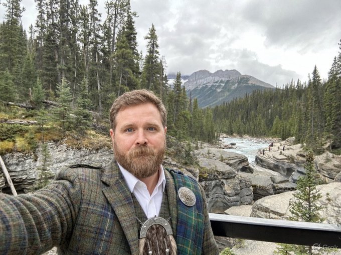 A bearded man in a plaid jacket and Scottish sporran takes a selfie with a mountainous landscape, evergreen forest, and turquoise river in the background, likely in the Canadian Rockies.