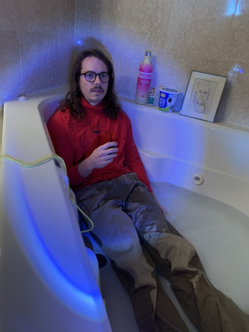 A man with long dark hair, glasses, and a mustache, wearing a red jacket and camouflage pants, sitting in a Jacuzzi tub illuminated with blue LED lights. Items including a Svedka bottle and a framed sketch are visible on the tub's edge.
