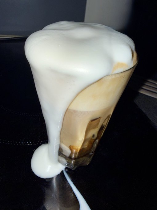 A close-up photograph of a glass containing a coffee beverage overflowing with thick white foam that spills dramatically down the side and pools onto the dark surface below.