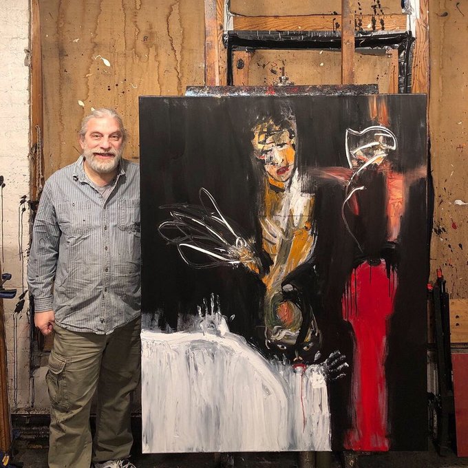 A gray-bearded man in a striped shirt stands next to a large, expressive expressionist painting featuring a figure holding a glass against a dark background with bold red and white elements.