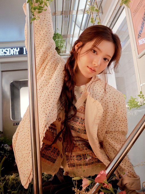 A young woman with a long braided hairstyle poses wearing a beige polka-dot blouse and a patterned knit vest in a greenhouse-like setting with plants.
