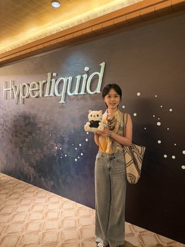 A woman wearing jeans and a t-shirt stands in front of a wall displaying the glowing 'Hyperliquid' logo while holding a small teddy bear and a striped bag.