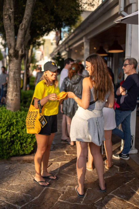 Two women chat outdoors, one in a yellow shirt and cap holding a branded yellow tote reading "BINANCE BLOCK CHAIN CRYPTO" alongside another woman in a gray dress, with a crowd mingling in the background of what appears to be a Binance-sponsored blockchain event.