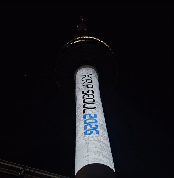 A tall illuminated tower at night displays 'EXPO 2026' vertically in white and blue text, viewed from a low angle looking up against the dark sky.