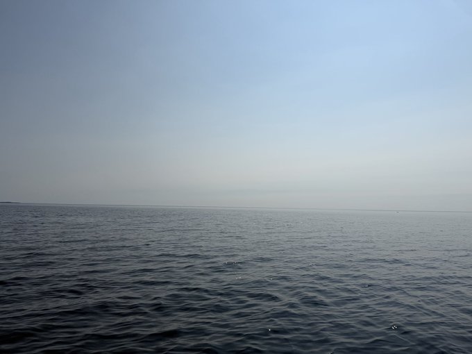 A serene photograph of a calm, dark blue body of water meeting a hazy, pale blue sky at the horizon with no land visible.