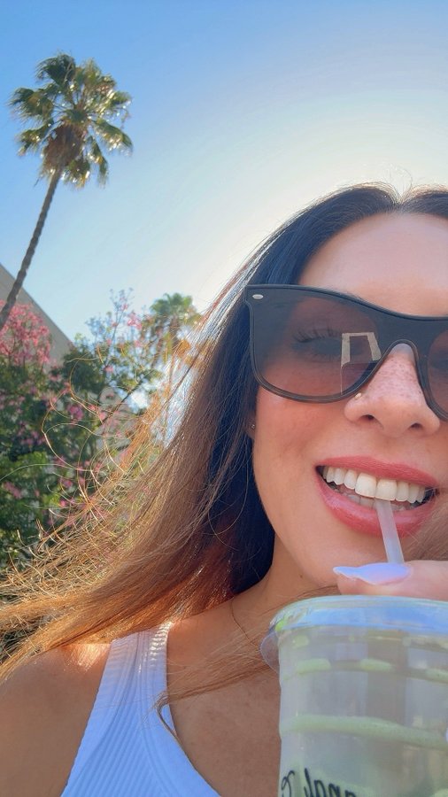 A selfie of a smiling woman wearing sunglasses, drinking from a cup with a straw outdoors with palm trees in the background.