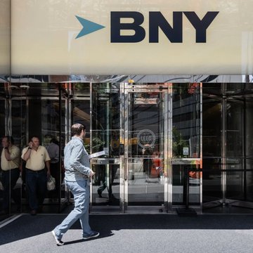 A man in a light blue suit walks past the glass entrance of a BNY (Bank of New York) building, with the company's blue arrow logo prominently displayed above the doors. The image illustrates the article's announcement about BNY, the world's largest custodian bank, expanding its services to include crypto staking.