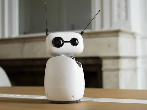 A small white robot with two black eyes and two antennae stands on a wooden table in front of a fireplace.