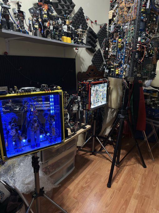 An electronics workshop or maker space filled with circuit boards, exposed wiring, and equipment, featuring a blue-lit illuminated case on a tripod displaying internal components and figurines.