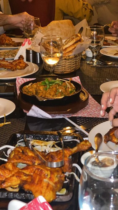 A dinner table is laden with various dishes including roasted chicken, a curry dish, and bread, with wine glasses and people's hands visible around the spread.