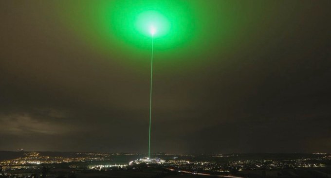 A bright green laser beam shoots up into the night sky above a cityscape, creating a glowing halo at its peak. The image pairs with a promotional post urging viewers to buy memecoins, using the laser's striking, attention-grabbing light as a visual metaphor for seizing a hot market opportunity.