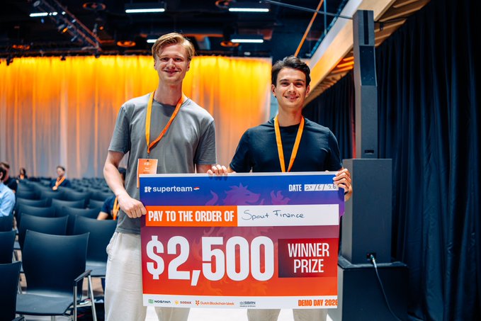 Two young men in lanyards holding a large '$2,500 WINNER PRIZE' novelty check from 'superteam' at what appears to be a conference event dated 'DEMO DAY 2024'.