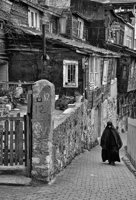 A black-and-white photograph of a narrow cobblestone street featuring old wooden buildings and stone walls, where a person wearing a long dark cloak and hat walks away from the camera.