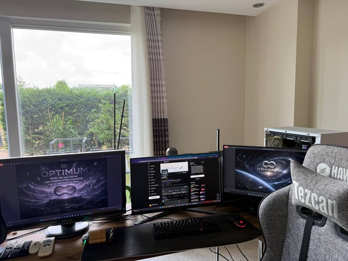 A modern home office setup with three gaming monitors displaying 'OPTIMUM' and 'OLYTHEIA' wallpapers, a black keyboard and mouse on a dark desk, a gray Tezcan gaming chair, and a view of greenery through a large window.