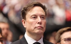 The image features a man dressed in a suit, standing in front of a crowd at an event or gathering. The man appears to be looking off into the distance with a serious expression on his face. The crowd around him consists of people who are also wearing suits and appear to be engaged in conversation or observing something happening nearby.