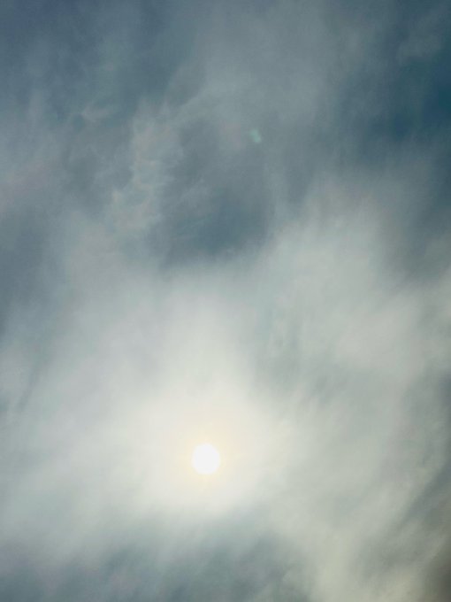 A photograph of the sun shining brightly through a layer of clouds in a blue sky.