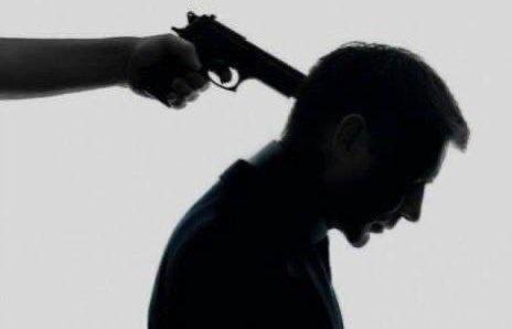 A silhouetted man in a dark suit holds his head down as another person's arm points a handgun at his temple.