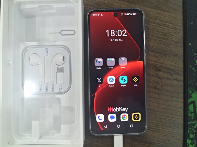 The image shows a WebKey smartphone (displaying the time 18:02 and a red-themed home screen with crypto apps like BingX and Bybit) alongside its packaging containing earphones, a charging cable, and a SIM tool. This relates to the post, where the user is reviewing/unboxing the WebKey phone—mentioning it costs $599, looks good, runs smoothly, and comes pre-installed with crypto trading apps—while also promoting the "Ave" project and offering to share a registration link for anyone interested.