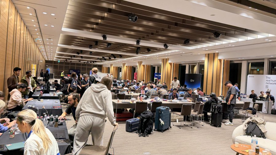 A crowded indoor lounge or co-working space with people working on laptops at long tables, surrounded by luggage, with banners mentioning 'Polkadot' and 'Humanode' visible.