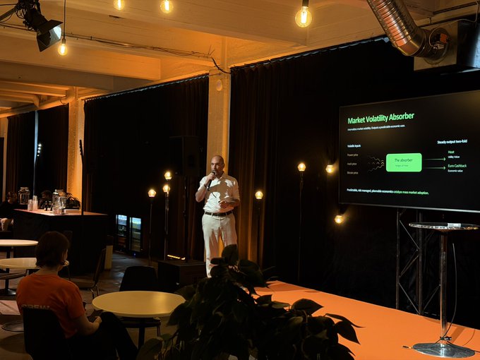 A man giving a presentation on stage in a dimly lit venue, speaking into a microphone while a screen behind him displays a slide titled 'Market Volatility Absorber'.
