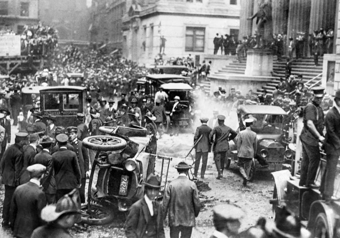 A black-and-white historical photograph showing a chaotic street scene with crowds, police officers, overturned vintage automobiles, and smoke in front of a large stone building with columns.