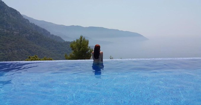 The image shows a woman standing at the edge of an outdoor swimming pool with her back turned towards the camera. The scene appears to be set in a mountainous area, as there are mountains visible in the background. The focus of the image seems to be on the woman and the pool rather than the surrounding landscape or other elements within the photo.