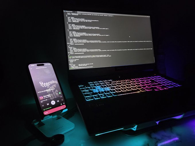 A laptop with a rainbow backlit keyboard displays code in a dark environment, next to a phone on a stand showing a music player titled 'Legends never die'.