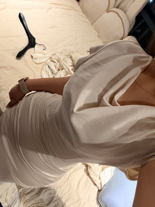 A woman wearing a beige satin dress and a smartwatch is sitting on a white bed with a black hanger and crumpled clothes nearby.