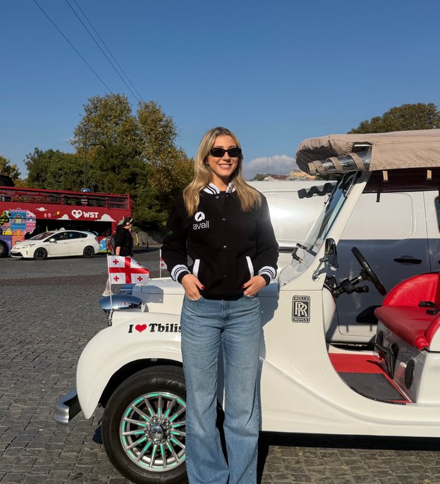 A woman stands smiling in front of a vintage white Rolls-Royce taxi in Tbilisi, wearing a black "Avail" hoodie, with an "I ❤ Tbilisi" decal and Georgian flag visible on the car. The image accompanies a post celebrating her one-year anniversary at Avail Project, captured during a rare break from work in the Georgian capital.