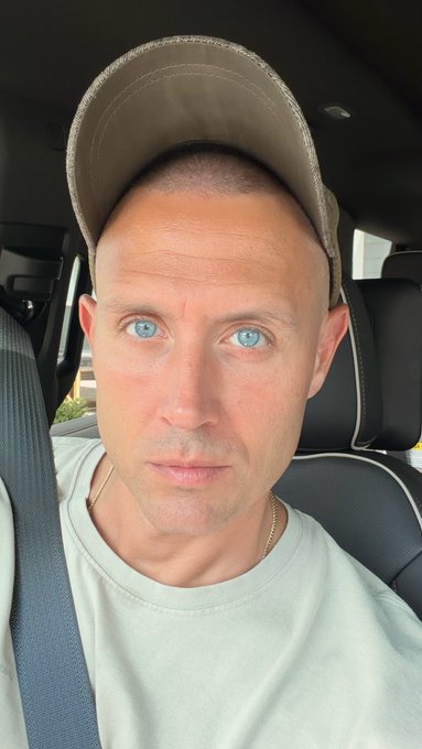 A close-up selfie taken inside a car shows a man with blue eyes and stubble, wearing a beige cap and a light-colored t-shirt with a gold chain necklace.