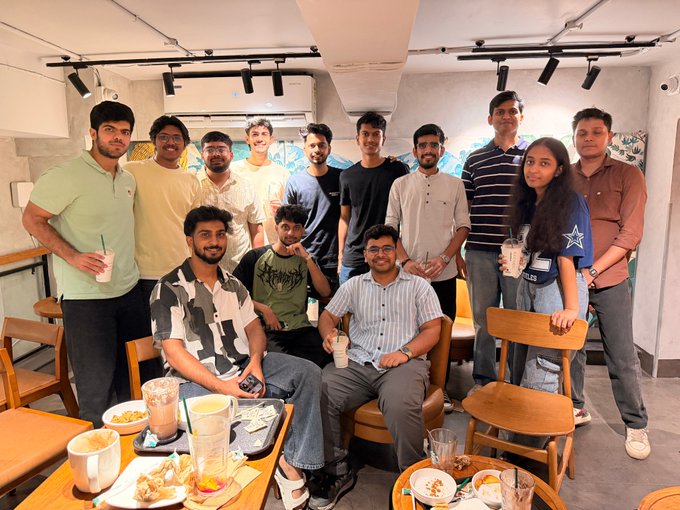 A group photo of approximately thirteen people posing together in a casual cafe setting, with food, drinks, and pastries on wooden tables in the foreground.