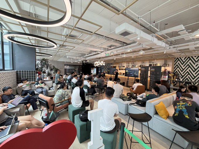 A spacious lounge or cafe area filled with people sitting on couches and chairs, with a presentation or screen visible in the background.