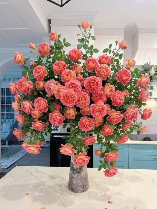 A large, lush bouquet of orange and pink garden roses arranged in a textured vase on a white marble kitchen island, with a light blue kitchen and tufted chair visible in the background.