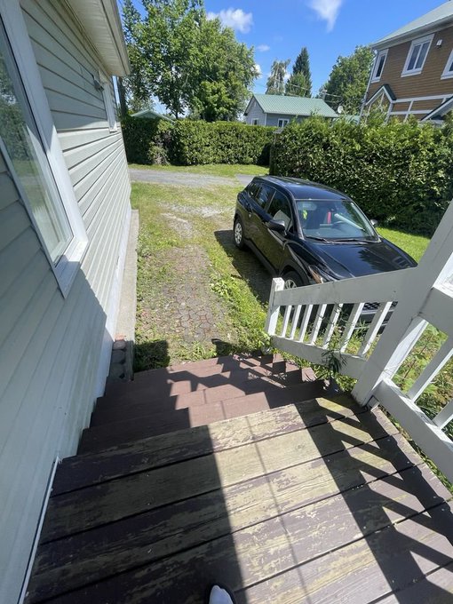 A view from wooden porch steps looking down at a black SUV parked in a grass driveway, with neighboring houses, green hedges, and trees visible under a blue sky with scattered clouds.