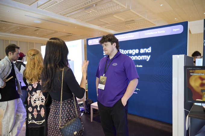A photo taken at a tech or crypto conference showing a young man in a purple 'W3S' polo shirt with a 'VIP' badge speaking with attendees in front of a banner reading 'Storage and Economy' with 'Compliance Ready' and 'lagoon' branding.