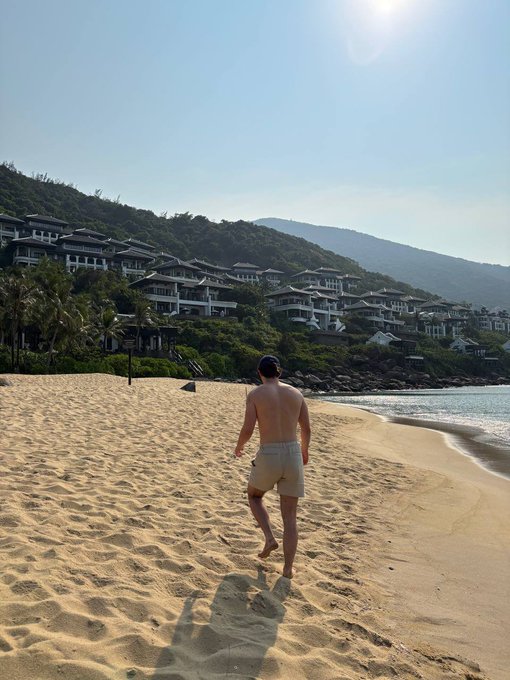 A shirtless man in beige shorts walks barefoot along a sandy beach toward a hillside resort with luxury villas overlooking the ocean.