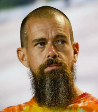 A close-up portrait of a bald man with a large bushy beard wearing a tie-dye orange and yellow shirt.