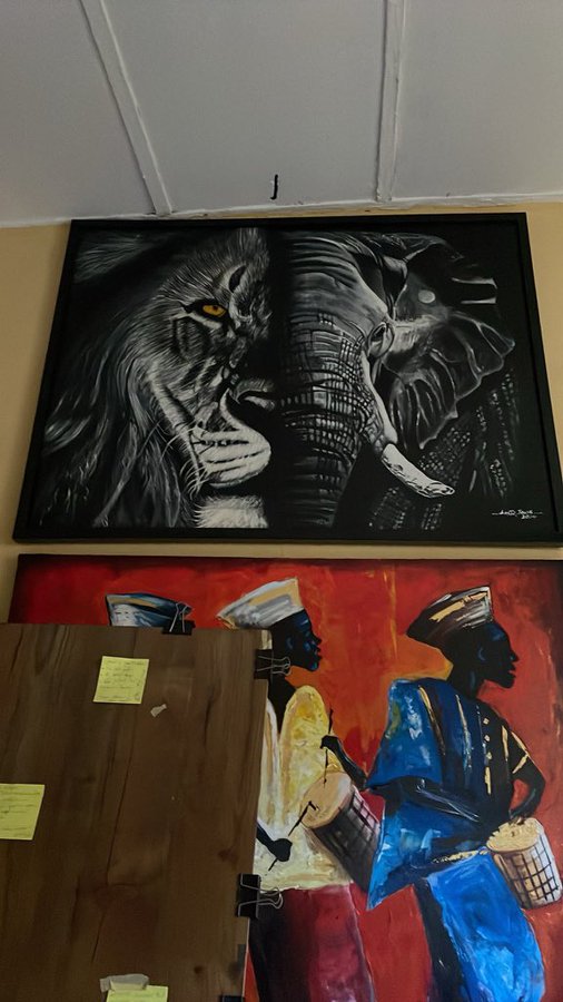 Two paintings hang on a wall: the top one depicts a lion and elephant in black and white, while the bottom shows figures playing drums in vibrant blue, red, and yellow tones.