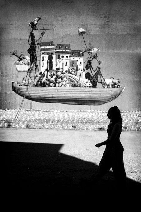 A black and white street art mural on a wall depicting a fantastical boat filled with houses and figures, with the silhouette of a woman walking past in the foreground.