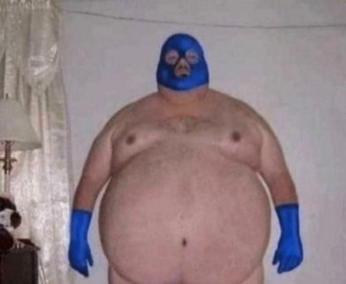 A heavyset person stands indoors wearing only a blue luchador-style mask and blue gloves.
