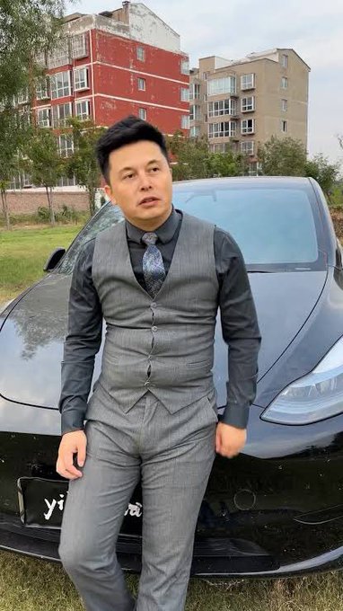A man in a gray vest and tie standing in front of a black Tesla car in an outdoor setting.