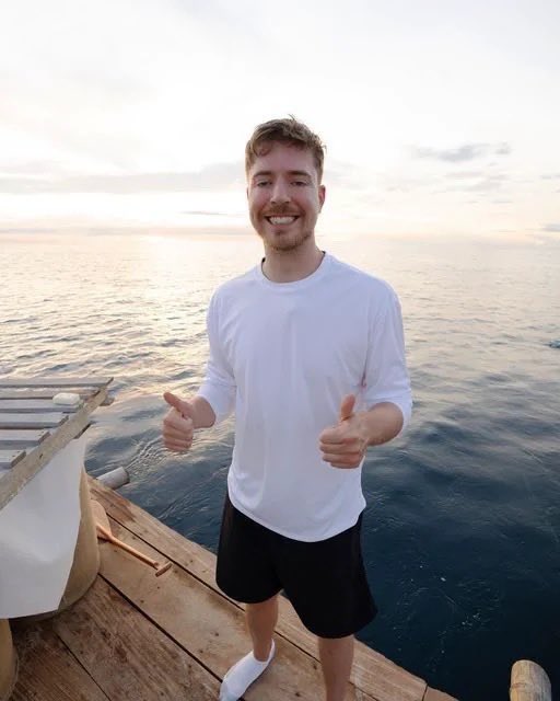 MrBeast stands smiling on a wooden deck by the ocean at sunset, giving two thumbs up while wearing a white long-sleeve shirt and black shorts.