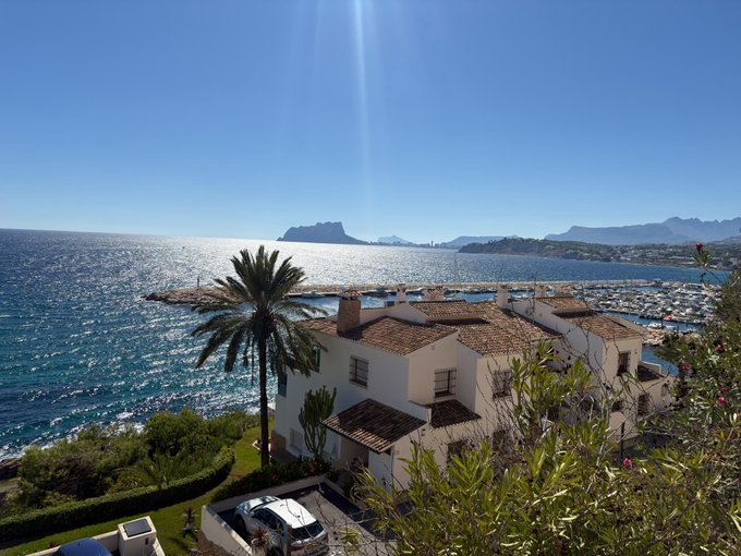 A sunny coastal view from a property, featuring a palm tree in the foreground, white Mediterranean-style buildings, a marina filled with boats, and mountains visible in the distance under a bright blue sky.