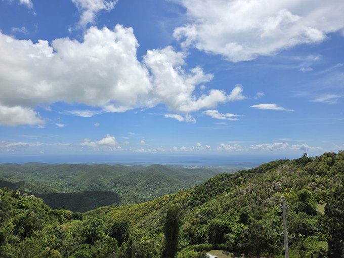 Rolling green hills and a partly cloudy sky stretch toward a distant coastline, offering a serene escape that fits the "touching grass" sentiment of stepping away from daily life. A utility pole and small structures in the foreground hint at human presence amid the lush landscape.