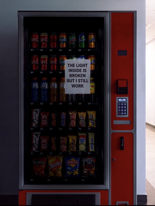 A vending machine with a hand-written sign taped to it reading 'THE LIGHT INSIDE IS BROKEN BUT I STILL WORK,' stocked with various drinks and snacks.