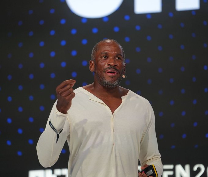 A man in a white button-up shirt speaks on stage with a headset mic, gesturing with his hand against a backdrop of blue lights and partial text reading 'EN 2'.