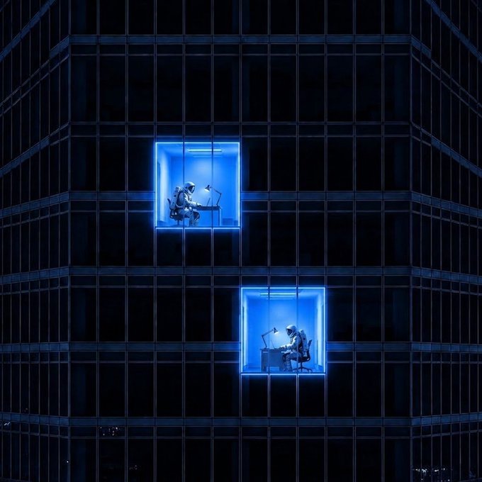 Two robot figures work at desks in illuminated blue office cubicles set within a dark glass skyscraper facade at night.